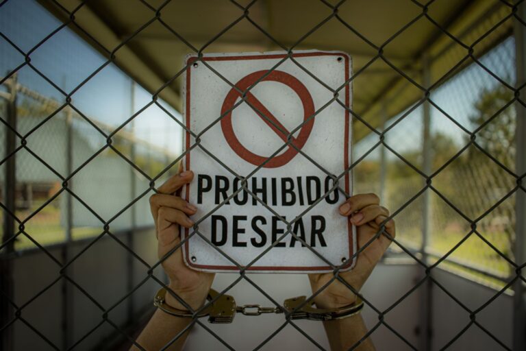 Handcuffed hands hold a 'Prohibido Desear' sign behind a chain-link fence, emphasizing restriction.
