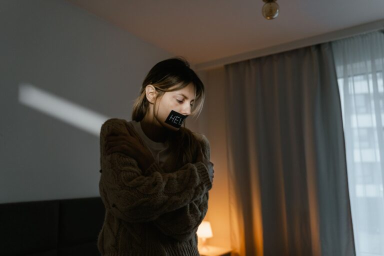 A young woman in a dim room wears a sweater and a help sign, expressing need.