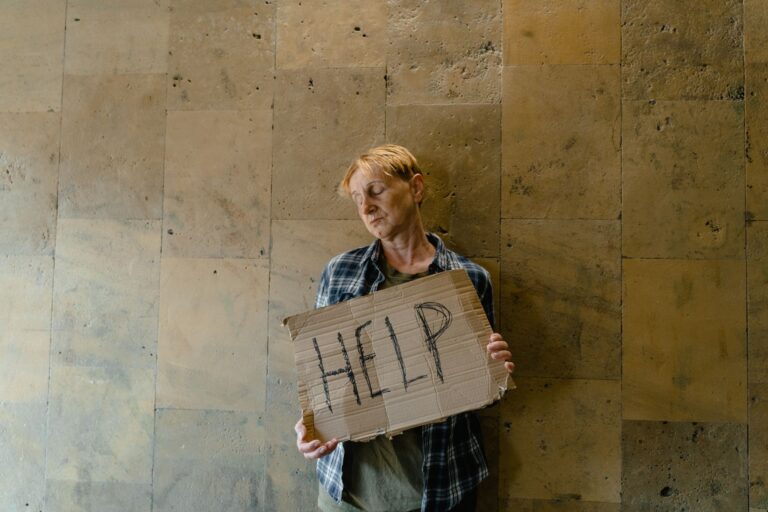 An elderly woman stands against a wall, eyes closed, holding a cardboard sign that reads 'HELP'.